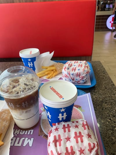 In Shtarkovo, Bulgaria, a close-up shot captures a fast-food meal on a table. In the foreground, a clear plastic cup filled with iced coffee with milk layers sits prominently. Beside it, a blue and white paper cup, branded with "HESBURGER" and a star logo, contains a pink beverage. To the right of that, another blue and white cup, similar to the one with the pink drink, is visible. Behind these cups, a blue tray holds a burger wrapped in white paper adorned with red "H" logos and stars, and a small portion of french fries in a paper wrapper. In the background, a red booth seat curves behind the table, and a glimpse of a restaurant interior with a person walking in the distance can be seen. The overall scene suggests a casual dining experience at a Hesburger outlet.