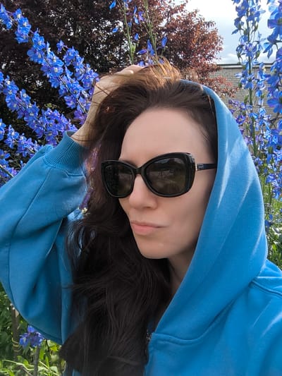 A woman with long, dark hair is captured in a medium shot, wearing a bright blue hooded sweatshirt and black sunglasses. She is looking off to her right, with her right hand resting on the back of her head, her fingers splayed through her hair. The background is filled with lush, tall blue delphinium flowers and some dark-leaved foliage. A brick building is visible in the upper right corner, partially obscured by the flowers. The lighting suggests it is daytime, possibly late afternoon, with the sun casting shadows and highlighting details. This scene is set in a garden in Vecumnieki, Latvia, characterized by the vibrant blue flowers and the natural setting.