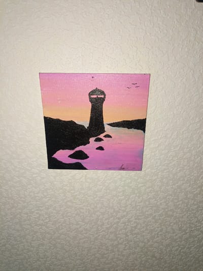 A small, square canvas painting of a lighthouse is mounted on a textured, off-white wall. The painting depicts a dark silhouette of a lighthouse against a gradient sky of pink and orange. Black landmasses and rocks are visible in the foreground, with water rendered in shades of pink and purple. Two small, dark bird silhouettes are in the upper right corner of the painting. The canvas is slightly tilted. The wall has a rough, uneven texture. There are no people or animals visible in the image. There is no information within the image that suggests a location in Magadan, Russia.