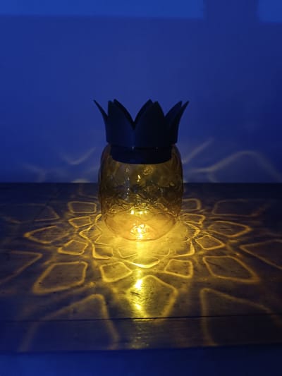 The image depicts a pineapple-shaped lantern placed on a dark surface. The lantern is yellow with a black, leafy crown. A light source inside the lantern projects a patterned, yellow glow onto the surface. The background is a dark blue, likely a wall. The location of the scene within Cherkovitsa, Bulgaria, is not discernible from the image. There are no people present in the image.