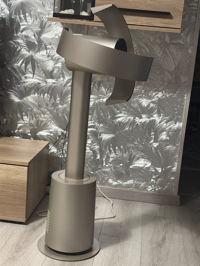An upright, silver bladeless fan stands on a light-colored wooden floor. The fan has a cylindrical base with a circular plate on the bottom. Above the base, a tall, slender column extends upwards, topped by a curved, loop-like head. The fan is positioned to the right of a light wood bedside table with a drawer. Behind the fan and table is a wall covered in patterned wallpaper featuring large, stylized leaves in shades of gray and white. The lighting in the scene is dim, with a spotlight illuminating the fan and the wallpaper. The image appears to have been taken indoors, likely in a bedroom or living area. There are no people or animals visible in the image. The specific location in Augsburg, Germany, cannot be determined from the visual information provided.