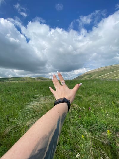 In the city of Andreevka, Russia, a person's arm and hand are outstretched towards a grassy landscape under a cloudy sky. The arm, featuring a tattoo on the lower portion, wears a black smartwatch. The hand, with long, white-tipped fingernails, displays a silver ring with a wavy design on the middle finger. The background consists of rolling green hills, interspersed with patches of darker clouds in the sky. The foreground is dominated by tall, wispy green grass and some small wildflowers, including purple and yellow varieties.