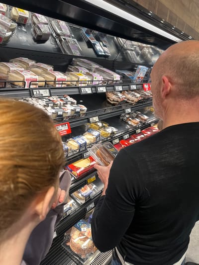 A man and a child are shopping for desserts in a supermarket aisle in Toormina, Australia. The man, bald with a beard, wears a black long-sleeved shirt and is holding a packaged dessert. The child, with reddish-brown hair, wears a grey top and is looking at the desserts. The aisle is stocked with various packaged cakes, cheesecakes, and tiramisu. A shelf below displays cheese products. Price tags are visible on the shelves. The lighting is bright, coming from overhead fluorescent lights.