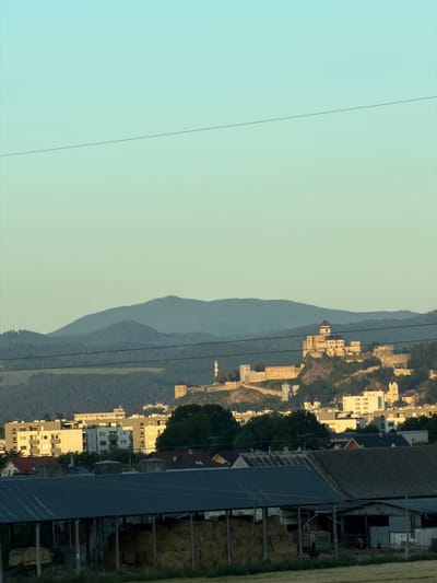 The image captures a panoramic view of the city of Trenčín, Slovakia, dominated by the imposing Trenčín Castle perched atop a hill in the background. The castle, a prominent historical landmark, features a large central tower and several connected structures. Below the castle, a collection of buildings, including apartment blocks and residential houses, form the urban landscape. The foreground is occupied by agricultural structures, such as sheds with dark roofs, and hay bales, suggesting a rural or semi-rural setting within or on the outskirts of the city. The sky is a clear gradient of light blue to pale yellow, indicative of either early morning or late afternoon light. Two thin dark lines, likely power lines, traverse the upper portion of the image.
