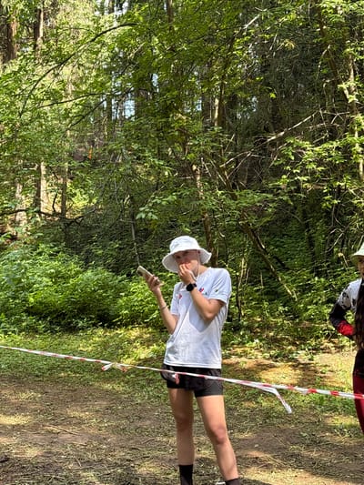 A person wearing a white bucket hat and a white t-shirt stands in a wooded area, holding a white cylindrical object in their right hand. They are wearing black shorts and black socks. A red and white striped ribbon or tape is visible in the foreground, stretching diagonally across the frame. The background is filled with lush green foliage and trees, suggesting a natural outdoor environment. Sunlight filters through the leaves, casting shadows on the ground. Another person, partially visible at the top of the frame, is wearing a dark shirt with red accents and a white bucket hat. The scene appears to be set during the daytime, possibly in a forest or park in Sergino, Russia. The mood seems relaxed and casual. No visible text is present on the person's clothing or the objects.