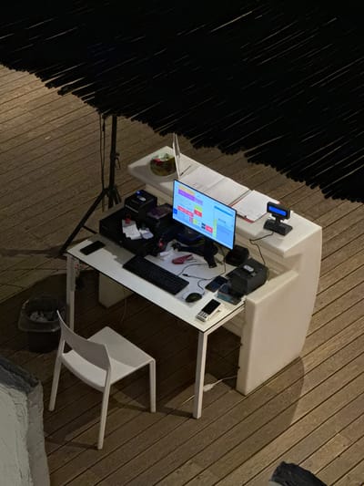 This is an overhead, medium shot of a reception desk set up outdoors on a wooden deck, likely in Capaci, Italy. A black, slatted awning provides partial shade overhead. The desk itself is white and L-shaped, with a computer monitor displaying a grid of colorful boxes, a keyboard, a mouse, a receipt printer, and several mobile phones on its surface. A small trash can and a white chair are situated to the left of the desk. The wooden deck planks run horizontally across the frame. The lighting suggests it is either dusk or nighttime.