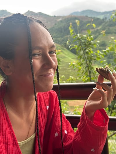 A young adult female is depicted outdoors, smiling and looking towards her right, in what is identified as Púng Luông, Vietnam. She has dark hair styled in numerous thin braids, some adorned with small silver-toned beads. A mole is visible above her right eyebrow and another near her right eye. She is wearing a textured, loose-fitting red garment with small white heart embroideries, over a ribbed, light olive green top. A delicate gold chain necklace is visible. In her right hand, she holds a partially consumed, dark brown, textured food item, possibly a wafer or biscuit, with a light-colored filling or topping. Her fingernails are long, manicured, and have a light, iridescent finish. She is positioned behind a dark brown or red railing, which extends horizontally across the mid-ground. The background consists of out-of-focus lush green foliage, trees, and rolling mountains, some showing signs of terraced cultivation, under a bright, overcast sky.