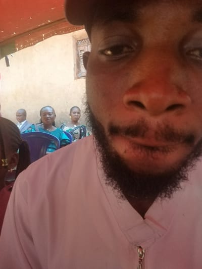 The image displays a gathering in Ochadam, Nigeria, with a dark-skinned male dominating the foreground, taking what appears to be a selfie. His face is positioned extremely close to the camera, revealing his right eye, nose, and a prominent beard covering his chin and jawline. He wears a light pink collared shirt with a silver zipper pull at the neckline. In the background, partially obscured, several individuals are seated. Visible from left to right are a person with dark hair, a bald male in a light suit jacket and white shirt, a female with dark hair in a teal-patterned top seated in a blue plastic chair, and another female in a patterned dress. Behind the seated individuals is a light-colored wall, possibly stucco, featuring a wooden window with horizontal slats. A red fabric canopy or awning is visible overhead.