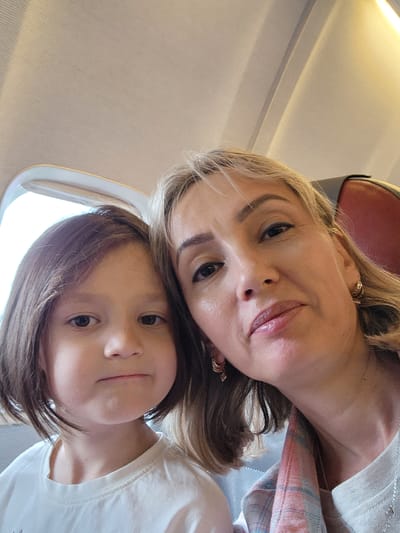 A selfie photograph shows two people: an adult female and a young girl, likely inside a passenger airplane. The girl is positioned above the woman's head, her face close to the camera. She has brown hair, fair skin, and dark eyes. The woman has blonde hair, light skin, and visible ear jewelry. A window is visible in the upper portion of the image, and airplane seating is visible in the lower portion. No information can be deduced about the location in the city of Stolbishche, Russia from the image.