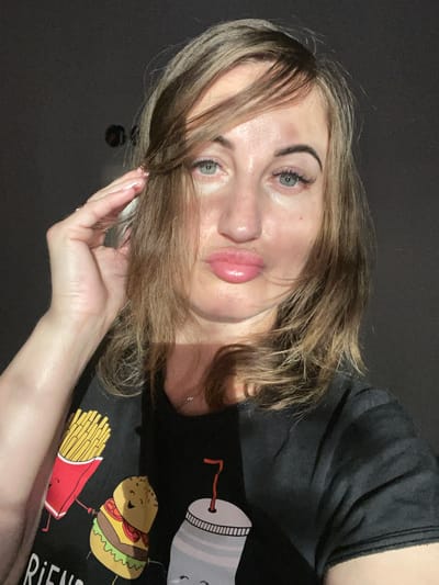 The image is a close-up selfie of a woman with light brown hair, blue eyes, and fair skin. She is wearing a black t-shirt with a graphic print of a hamburger and french fries. Her left hand is raised to her head, and she is puckering her lips. The background is dark and appears to be an indoor setting. There are no identifiable landmarks or context that would suggest a specific location in Zarichne, Ukraine. The image focuses on the woman's face and upper body.