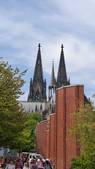 The image shows the Cologne Cathedral, a UNESCO World Heritage Site, in Cologne, Germany. The cathedral, with its iconic twin spires, dominates the background. In the foreground, a group of people are walking along a brick-lined pathway. The pathway is bordered by lush green trees on the left and a red brick building on the right. The sky is a pale blue with scattered clouds. The overall scene depicts a sunny day with tourists enjoying the sights of the historic city.