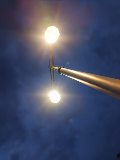 The image displays a low-angle perspective of a street light assembly against a dark blue, cloud-dotted sky in Cianjur, Indonesia. A metallic street light pole ascends diagonally from the lower right toward the center of the frame. Two thinner, perpendicular arms extend from this main pole, each supporting a bright, polygonal-shaped light fixture. One illuminated fixture is positioned in the upper left quadrant, and the other is located in the lower-central left. Both lights emit strong, warm-toned illumination, generating visible lens flares. The environment is dark, indicating evening or night, with no individuals or discernible activity present.