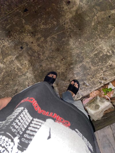 The image shows a person's feet wearing black slides, standing on a wet, textured concrete surface. The person is wearing dark pants and a black t-shirt with red and white text. Some debris, including leaves and a piece of pink insulation, is visible on the ground to the right. The scene appears to be outdoors, possibly a street or walkway in Banjarmasin, Indonesia, indicated by the presence of wet concrete and natural elements.