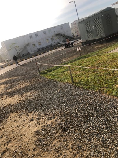 The image depicts an outdoor scene in النعيلة, Saudi Arabia, characterized by modular residential or office structures under a bright, hazy sky. In the foreground, a path of gravel and sand transitions into a strip of green grass, separated by a low barrier of red and white striped tape secured by wooden posts.
In the midground, a paved or graveled roadway bordered by yellow and black curbstones is visible. A male individual, dressed in a dark jacket and light-colored trousers, walks from left to right on this roadway. Further along the roadway, near the right side of the frame, three individuals are present: one stands wearing a light top and dark pants, while two others crouch or sit on the ground, engaged in an unclear interaction. Adjacent to the roadway on the right, a large, dark gray utility box or generator, equipped with vents and a warning decal, stands on a raised platform. A tall street light post rises above this structure.
In the background, multiple two-story white modular buildings, resembling prefabricated units, line the roadway. These structures feature numerous windows, external staircases, and visible air conditioning units mounted along their lower facades. Sparse young trees are planted alongside the buildings. More similar structures are visible further in the distance, suggesting a larger complex or camp. The overall lighting indicates daytime conditions with strong sunlight.