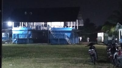 The image shows a nighttime scene in Uteunreutoh, Indonesia, likely a gathering place with a lawn and some buildings. In the foreground, several motorcycles are parked on the grass. The main building is elevated and painted blue and white, with a dark roof and lights illuminating the interior, where people can be seen. Stairs lead up to this building. To the right, a smaller structure with a pitched roof is visible, and another building is partially seen on the far left. A person in light-colored clothing is walking away from the camera in the middle ground, towards a lit doorway. The environment appears to be outdoors with some vegetation.