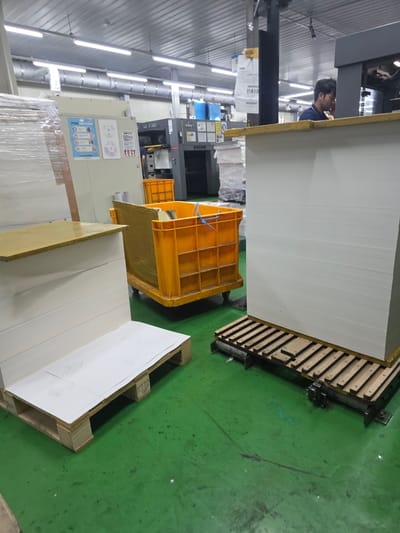 The image depicts an indoor scene, likely a printing or manufacturing facility in Soseok-ri, South Korea. The environment is utilitarian, with a green, epoxy-coated concrete floor and a suspended ceiling with fluorescent lighting. In the foreground, a large stack of white paper sits on a wooden pallet, which is itself on a metal track system. To the left, another stack of paper is partially visible, also on a pallet. A large, orange plastic bin with visible contents and yellow plastic wrapping is positioned in the mid-ground. Behind this bin, a printing press or large industrial machine is visible, labeled "RMGT". A man, wearing a dark blue shirt with a light blue collar, is standing at the machine, appearing to be engaged with its operation. He has dark hair and is looking down. To the left of the machine, a beige industrial cabinet with several informational posters is mounted on the wall. The background shows more of the facility, with various pipes, machinery, and stacked materials. The overall impression is of an active workspace involved in paper processing or printing.