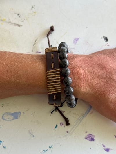 The image shows two bracelets on a person's wrist. One bracelet is made of dark brown leather with lighter brown string wrapped around it, featuring two stitched eyelets. The second bracelet, worn closer to the hand, is composed of dark gray, spherical beads. The wrist is placed on a white surface with faint smudges of blue, purple, and black paint. The location in Tam Kỳ, Vietnam, is not discernible from the image.