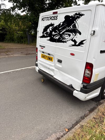 The image captures the rear of a white Ford Transit van, parked on the side of an asphalt road with a grassy verge, in Hazlemere, United Kingdom. The van exhibits visible damage, primarily to its rear bumper and bodywork.

The main subject is the white van, which has a large, intricate black vinyl decal on its left rear door, depicting a motocross rider on a dirt bike in mid-air. The word "MOTOCROSS" is written vertically as part of this decal. Below the right rear door, the word "TRANSIT" is visible, along with a red "Ford" oval logo. The yellow UK number plate, "GA57 DWA", is centrally located on the lower rear door.

Notable damage includes a significant dent and creasing on the right side of the rear bumper, extending upwards onto the white bodywork near the right rear light, where a crack is also apparent. A small, green and yellow decorative object, possibly a toy or charm, hangs below the damaged right side of the bumper.

The scene takes place during daylight hours, with no specific weather conditions evident beyond a dry road. Trees and foliage are visible in the background, suggesting a leafy, possibly residential or semi-rural setting. The van appears stationary, with no people visible.