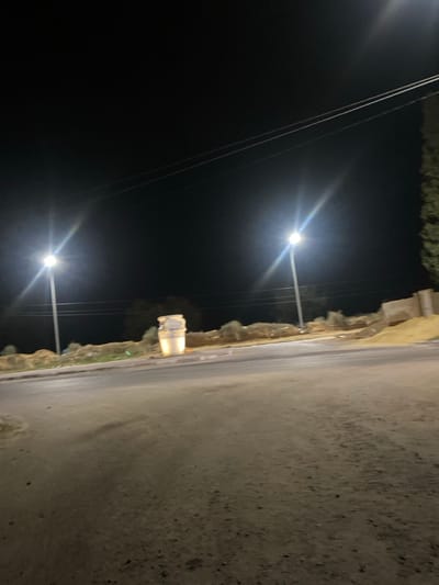 This nighttime image depicts a road in الغنادة, Tunisia. Two prominent streetlights on poles illuminate the scene, one positioned on the left and another further right of center, both emitting bright white light. Overhead utility lines are visible spanning horizontally across the upper third of the frame. In the mid-ground, a large, light-colored decorative urn or vase stands on a raised, vegetated median, illuminated from below or by ambient light. The foreground consists of a dark, unpaved or dirt road surface, with a paved section and curb line visible on the left. Beyond the median, a low, vegetated embankment with sparse green and dry shrubbery extends across the background. To the far right, a portion of a tall coniferous tree and a low, light-colored concrete wall are visible on an elevated section. No individuals or vehicles are present.