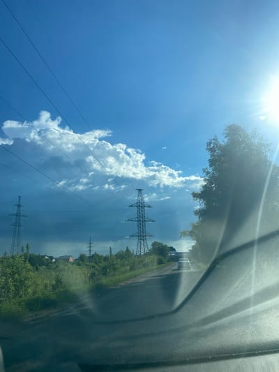 The image shows a view from inside a vehicle on a paved road, looking forward and slightly to the right. The road curves to the right and leads towards a white car in the distance. On the left side of the road, there is lush green vegetation and several tall power line towers. Beyond the vegetation, a few houses are visible. The sky is a bright blue with scattered white and dark clouds, and the sun is shining intensely on the right side of the frame, causing lens flare and obscuring details of the trees in that area. The overall scene suggests a rural or suburban environment.