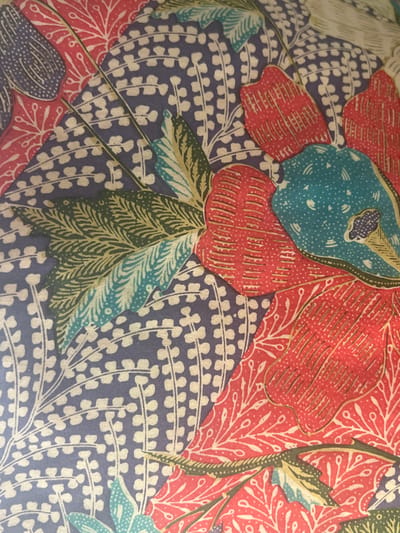 The image shows a textile pattern with floral and abstract motifs. Large red petals dominate, adorned with fine parallel lines and small tendrils. Teal-colored elements appear with dotted patterns. The background is a muted lavender or purple color filled with a repeating motif of small, off-white semi-circles arranged in rows. Green and teal leaf-like shapes are scattered throughout. The overall impression is a vibrant and intricate batik-style design.