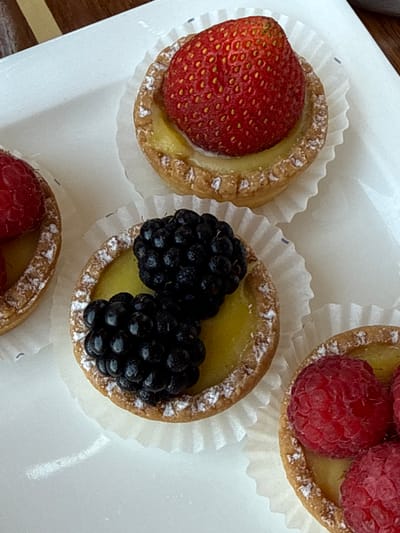 Four fruit tarts are arranged on a white rectangular plate. From top to bottom and left to right, the tarts feature a strawberry, two blackberries, and three raspberries as toppings. Each tart has a golden-brown crust and a yellow filling. The tarts are presented in white paper liners and are dusted with powdered sugar. The plate is positioned on a wooden surface, possibly a table. The scene is indoors, and the lighting suggests a daytime setting. There are no people visible in the image. The location of Vouliagmeni Municipal Unit, Greece is not discernible from the image alone.