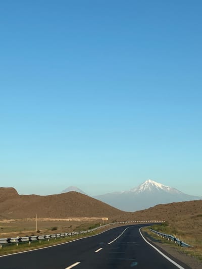 The image shows a highway curving through a barren, hilly landscape with mountains in the distance. The sky is clear and blue. In the foreground, a black asphalt road is bordered by a guardrail. The hills are covered in dry grass and scrub. In the background, two mountains are visible, one of which is snow-capped. Based on the visual cues, this scene is likely Mount Aragats, a prominent mountain in Armenia, and the highway is likely a road leading to or from it. Given Zangakatun's proximity to Mount Aragats, this scene could be located in the vicinity of the city. There are no people or animals visible in the image.