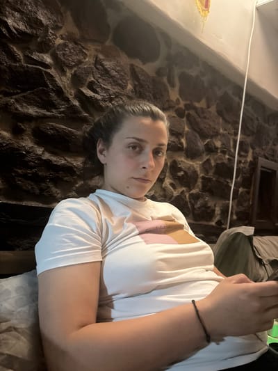 A woman is centrally featured in an indoor setting, positioned on the right side of the frame and captured from the chest up. She has dark hair pulled back and looks directly at the viewer with a calm, neutral expression; a small mole is visible on her left cheek. She wears a white, short-sleeved t-shirt, beneath which a pink and light brown garment with faint, light-colored text is visible on her right shoulder area, appearing to read "first rule." A thin, dark bracelet is on her right wrist. Her left arm is bent with her hand near her chest, while her right arm rests on a light-colored, patterned surface, likely upholstery.
The background is dominated by a dark, rustic stone wall, composed of irregularly shaped dark grey and brown stones with lighter mortar lines. In the upper left, a white ledge or shelf holds a small, yellow and red decorative item. A thin white cable runs horizontally across the upper wall. To the upper right, a dark, rectangular opening, possibly a window or niche, and some draped olive green or dark fabric are visible.
The scene depicts the woman posing for a photograph. The lighting is dim and diffused, suggesting an indoor evening environment. The rustic stone wall and potential traditional decor hint at a characteristic interior in Belopoptsi, Bulgaria.