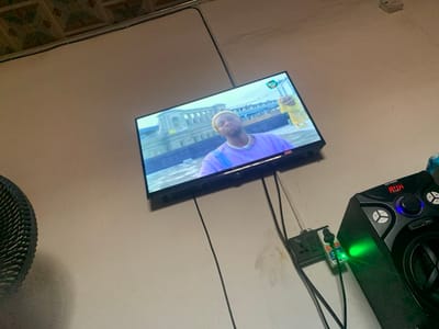 A television is mounted on a white wall displaying a man wearing a purple shirt and a silver chain, holding a yellow bottle. Below the TV, electrical cords run to a power strip. A black speaker with multiple buttons and a green light is positioned to the right of the power strip. The room has a visible ceiling with a patterned tile design and an electric fan is partially visible on the left side.