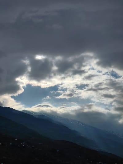 This is a vertical, low-angle photograph of a dramatic sky over the mountains near Gondhla, India. The sky is filled with dark, ominous storm clouds in the upper portion of the frame, but a large opening reveals bright blue sky with a patch of sunlight shining through. White, fluffy clouds are visible both above and below the opening in the dark clouds.
The mountain range in the foreground is dark and shadowy, with some areas of snow or ice visible on the upper slopes. The terrain slopes downwards from the upper left to the lower right of the frame. Small structures, possibly tents or buildings, are scattered on the lower slopes of the mountain. The overall impression is one of rugged, natural beauty and the power of the elements.
