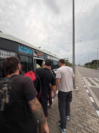 In Hyderabad, India, under a cloudy sky, a group of men are boarding an AC COACH bus with a "Welcome" sign. The bus, with text in blue and orange, has the number "7780641655" and "440569..." visible on its side. The men, dressed in casual attire, are walking along a brick-paved sidewalk towards the bus entrance. One man, in the foreground, has a backpack and tattoos on his arm. The scene is set along a road with streetlights and a pedestrian crossing.