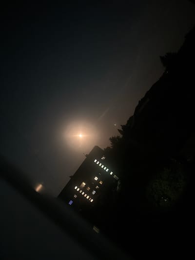 The image depicts a nighttime scene in Peremyshliany, Ukraine, characterized by a bright moon or other light source illuminating the sky and casting a soft glow. A multi-story building, angled diagonally in the frame, is visible with several windows lit from within, suggesting occupancy. The surrounding area appears dark, with silhouettes of trees and possibly hills or elevated terrain to the right. Power lines are also present, crossing the sky. The overall atmosphere is somewhat hazy, possibly due to atmospheric conditions or the light source itself. The perspective suggests the photo was taken from a slightly elevated position looking up and across the scene.