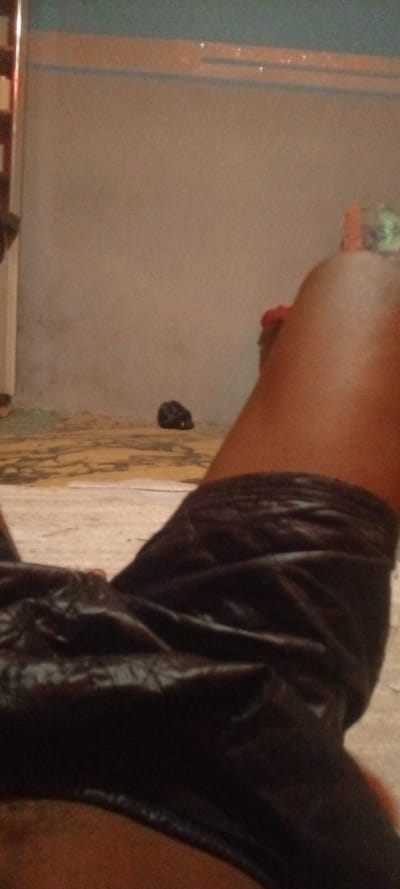 The image presents an indoor scene from a first-person perspective, looking down at a person's legs while they are resting or reclining. The subject's bare legs, which have a dark brown skin tone, are visible, extending into a pair of dark, possibly black, shorts. The shorts appear to be made of a soft, slightly textured or velvety material.
In the background, a light-colored floor with abstract, darker patterns is visible, suggesting either patterned tiles or a rug. Further back, a plain, light grey or beige wall rises, topped by a darker horizontal trim line. Above this trim, the upper section of the wall appears in a muted blue-green hue. Several small, faint, and indistinct reflective marks, which in close-up resemble symbols or inverted question marks, are sparsely visible on this upper wall area. On the floor, closer to the main wall, a small, dark, unidentifiable object is present.
The lighting in the scene is dim and warm-toned, suggesting low natural light or artificial illumination, possibly during the evening or night. The overall impression is one of casual relaxation. The location context is Ago-Abo, Nigeria.