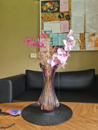 In a room with a textured olive-green wall, a black leather sofa is visible. On a wooden table in front of the sofa, a purple glass vase holds a bouquet of pink orchids and dried flowers. To the left of the vase, a black pair of glasses lies on the table next to a stack of three colorful cards. Behind the sofa, a bulletin board displays various papers, including a colorful floral picture at the top. A white electrical outlet with two sockets is mounted on the wall to the left of the bulletin board. The scene appears to be in a domestic or office setting in Molėtai, Lithuania.