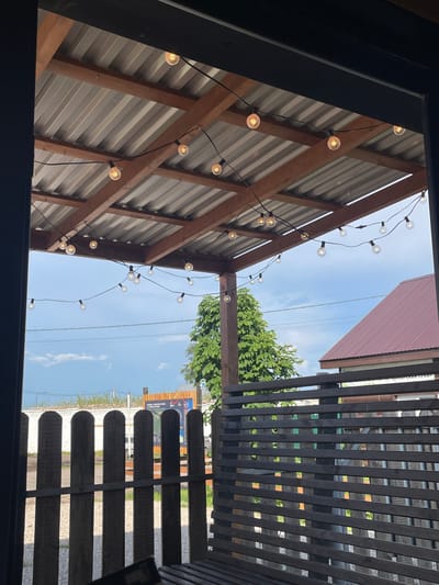 The image shows an outdoor area with a covered patio. The patio has a corrugated metal roof supported by wooden beams. A string of incandescent bulbs is strung across the underside of the roof. Through the opening of the patio, a wooden fence with vertical planks is visible, followed by a darker horizontal slatted fence. Beyond the fences, a large green tree stands to the left, and a building with a reddish-brown, corrugated roof is seen to the right. The sky is cloudy. In the background, there appears to be some kind of informational signage or display. The scene is consistent with an outdoor seating area at a restaurant or cafe in Shiryaevo, Russia, likely during daylight hours. There are no people or animals visible in the image.
