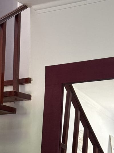 The image shows an interior scene featuring a wooden staircase and a wall with white paint. The staircase has dark brown wooden railings and steps. One section of the staircase appears to be under repair or damaged, with a splintered edge on a horizontal support. A large, dark red-framed mirror or doorway is positioned in the foreground, partially obscuring the view of the staircase. The reflection in the mirror shows more of the staircase and a white wall with decorative molding at the ceiling. The scene is well-lit, possibly by natural light coming from an unseen source. The location is identified as a residential building in Oeiras, Portugal. There are no people visible in the image.