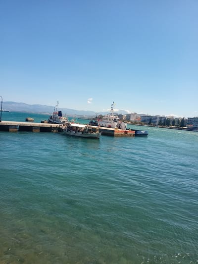 The image depicts a sunny day scene at the port of Anthidon, Greece. In the foreground, the clear turquoise water of the sea is visible, with some sunlight glinting off the surface and revealing the sandy bottom below. A concrete pier extends into the water from the left, supported by visible pillars. Moored to the pier are several boats, including a white and blue tugboat and a smaller fishing vessel with a white canopy and numerous people on board. In the background, a line of buildings stretches along the coastline, interspersed with trees and mountains in the distance under a clear blue sky. The overall atmosphere is tranquil and depicts a typical day at a Greek port.