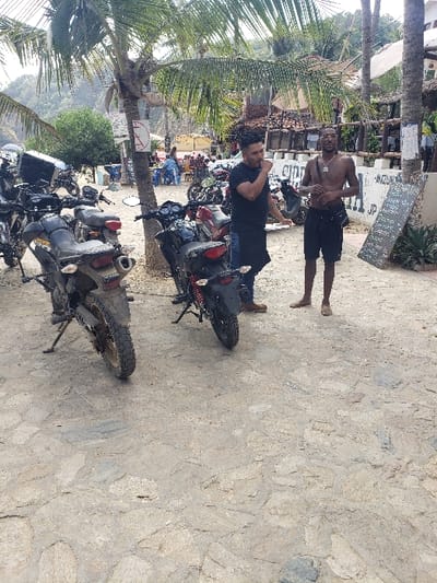 A man in a black shirt sits atop a motorcycle, while another man, shirtless, stands nearby. Several other motorcycles are parked in a row. The ground is paved with stones, and palm trees stand in the background. A sign is visible in the distance. The overall scene appears to be outdoors during the daytime.