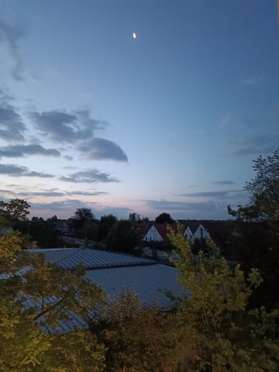 A twilight view of a suburban landscape in Germering, Germany, shows a clear sky with a crescent moon high above. Wispy clouds streak across the upper portion of the sky, transitioning to softer hues near the horizon where a faint pinkish glow suggests the last vestiges of sunset.
Below the sky, a collection of houses with red-tiled roofs are nestled amongst lush green trees. The buildings appear to be single-family homes, with several visible white facades. In the foreground, two large, leafy trees frame the scene, their branches extending towards the center. Behind the trees, a large, corrugated metal roof of what appears to be a commercial or industrial building dominates the lower part of the image. The overall impression is one of a peaceful evening in a residential area.