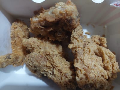A close-up, overhead shot captures several pieces of golden-brown fried chicken arranged in a white and pink striped cardboard container. The chicken pieces, appearing to be wings and possibly drumettes, have a crispy, textured coating. Some pieces are piled on top of others, creating depth in the image. The lighting is bright, suggesting it might be daytime, and the focus is sharp on the chicken, highlighting its appetizing appearance. There is no visible text in the image, and the background is a soft blur of the container's interior. The setting appears to be indoors, possibly at a fast-food establishment or a home. The overall impression is one of a ready-to-eat meal.