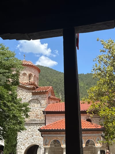The image shows a partial view of the exterior of the Bachkovo Monastery in Bachkovo, Bulgaria, from a low angle, framed by wooden structural elements. A section of the stone and brick monastery with a red-tiled roof is visible. The building features arched windows and decorative paintings on the exterior walls. Green foliage, including trees, partially obscures the view. In the background, a mountain covered in greenery rises behind the monastery. The sky is blue with scattered white clouds.