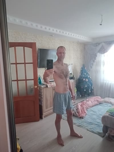 A middle-aged Caucasian man with a shaved head and chest hair is standing in what appears to be a bedroom in Suklieia, Moldova. He is wearing blue patterned boxer shorts and is holding a black smartphone in his right hand, taking a selfie in a large mirror. The room is decorated with a Christmas tree, a dresser with an iron on it, and a television. Curtains are visible behind the Christmas tree, and a window allows light to enter the room. There are also blankets and pillows on the floor, suggesting a living or sleeping area. The man's left arm is partially visible, and his bare feet are on the light-colored floor. The overall impression is of a casual, personal moment captured in a home setting.
