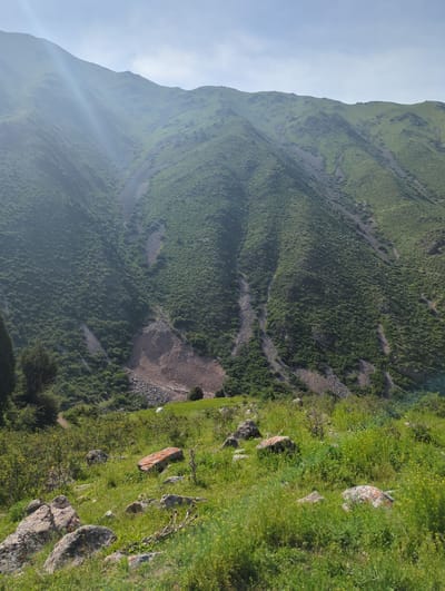 The image shows a mountain landscape in Chunkurchak, Kyrgyzstan. The mountain is covered in green vegetation, interspersed with rocky areas and trails. The sky is partly cloudy. The foreground features rocks and grass.