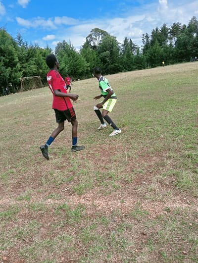 The image depicts an outdoor soccer match or practice taking place in Lwandeti ward, Kenya. The scene is set on an uneven, partially dry grassy field under a blue sky with scattered white clouds. In the foreground, two youthful males with dark complexions are actively engaged. The individual on the left wears a red short-sleeved jersey with black details, black shorts, dark socks, and black athletic shoes, standing with his body angled right and head turned left, looking upwards. The individual on the right wears a lime green short-sleeved jersey with black trim, black shorts, black socks, and white athletic shoes, positioned in a low, crouched, defensive posture, facing the first player. A white-framed soccer goal with a net is visible in the mid-ground on the left side of the field. Several other individuals, presumably players, are present further in the background, including a figure in red near the goal and a yellow figure on the distant field. A dense line of tall green trees forms the horizon, suggesting a natural or rural setting.