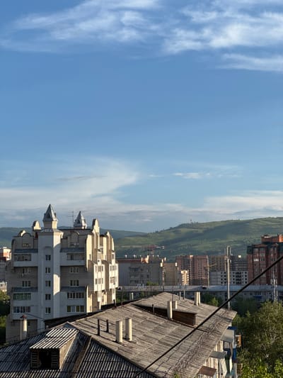 This image, captured from a high vantage point in Chita, Russia, depicts a sprawling urban landscape under a bright blue sky with scattered white clouds. The foreground is dominated by the weathered, grey, corrugated roofs of older buildings, complete with chimneys and what appears to be a small dormer window with its shutter ajar.
In the midground, a striking, multi-story building with a distinct, almost castle-like architectural style stands out. It features white and light beige brickwork, pointed turrets topped with grey caps, and numerous windows arranged in a rhythmic pattern. Behind this building, a dense collection of residential and commercial structures stretches across the cityscape, varying in height and architectural design, with some showing warm brick facades and others concrete exteriors. Electrical wires crisscross the scene, suggesting a developed urban infrastructure.
The background is characterized by rolling, green hills, indicating the natural topography surrounding Chita. The sunlight casts shadows, highlighting the textures of the buildings and the contours of the landscape. The scene conveys a sense of a typical day in this Siberian city, with a mix of older and more modern architecture set against a backdrop of natural beauty.