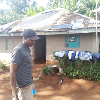 The image displays an outdoor scene in Getenga ward, Kenya. A single adult male is positioned in the left foreground, oriented to his right with his head slightly bowed. He wears a black baseball cap, a grey short-sleeved polo shirt, and light-colored trousers, holding a small blue container in his left hand. Behind him stands a single-story house with light-colored walls, likely plaster over an earthen structure, and a weathered, corrugated metal roof. Two small solar panels are mounted on the roof slightly off-center to the right, with a white garment draped over the roof adjacent to them. A small, light-colored wooden window frame is visible on the house's left side. A dark, open doorway is centrally located on the house's facade. To the right of the doorway, a teal-colored metal box with a small reflective square is affixed to the wall, with wires extending from it. Green bushes line the base of the house in the mid-ground, over which various items of clothing are draped for drying. On the reddish-brown soil in front of the bushes, a metal basin and a single dark-colored shoe are visible. The background consists of lush green, large-leafed trees, indicative of banana plants. The scene is illuminated by strong daylight.