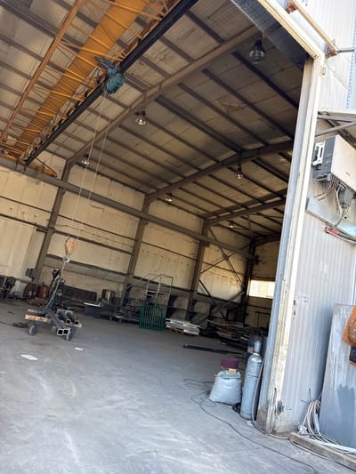 The image shows the interior of an industrial warehouse in Tsaratsovo, Bulgaria. The warehouse has a high ceiling with exposed metal beams and corrugated metal roofing. A large overhead crane is visible, running along a track on the ceiling. Various industrial equipment and materials are scattered throughout the space, including metal pipes, a welding machine, gas cylinders, and a cart with metal components. The floor is concrete. The scene appears to be a workspace, possibly for metal fabrication or construction. The lighting is a mix of natural light coming from the open doorway and artificial lights mounted on the ceiling.