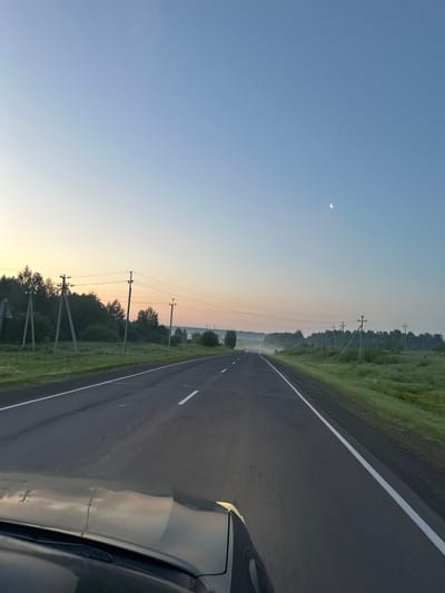 The image shows a view from the driver's seat of a car traveling on a paved road. The road has white dashed lines marking the lanes. The landscape on either side of the road is rural, with green grass and trees. There are utility poles with power lines running along the road. In the distance, there is some fog or mist. The sky is a clear, light blue, with a crescent moon visible. The sun appears to be rising or setting, as evidenced by the soft, warm light on the horizon. The car's hood and windshield wipers are visible in the foreground, reflecting the sky and landscape. The scene is peaceful and serene, suggesting a journey in the early morning or late evening. The location is specified as Krapivinskiy, Russia.