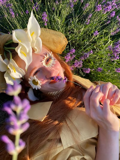 The media file shows a close-up, overhead view of a person lying in a field of lavender. The person has long, reddish-brown hair and is adorned with flowers. White calla lilies are arranged around the person's head, and daisy-like flowers are placed on their eyelids. Sprigs of lavender are also present, with one resting on the person's nose and others blurring in the foreground. The person is wearing a light-colored garment, possibly a dress or shirt, with a textured fabric. Their hands, with manicured nails, are visible near their face, interlaced. The scene is bathed in warm sunlight, suggesting it is daytime, likely late afternoon or early evening due to the soft lighting and shadows. The abundance of lavender and the natural setting indicate the location is outdoors, in a rural or agricultural area, possibly a lavender farm. The overall impression is one of tranquility and natural beauty, with a hint of artistic or surreal expression due to the floral decorations on the face. No text is visible in the media file. The context provided places the scene in Verkhneye Uchdere, Russia.