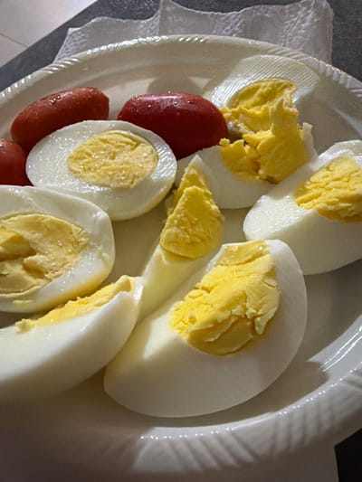 A close-up shot shows several hard-boiled eggs, some cut in half, and a few small, elongated red objects, possibly cherry tomatoes, on a white plate. The eggs display distinct white and yellow portions. The plate has a textured rim. The background appears to be a dark surface, possibly a countertop, with a white paper napkin peeking into the frame. The lighting casts shadows on the plate and highlights the glossy surface of the eggs. The scene is indoors, likely in a kitchen or dining area. The image does not contain information about the city of Gedera, Israel.